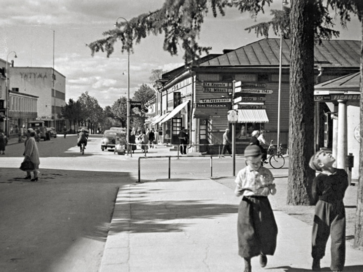 Lappeenrannan Kauppakadun ja Valtakadun risteys 1950-luvulla, etualalla oikealla kaksi poikaa kävelee jalkakäytävällä. Taustalla näkyy vanhoja liikerakennuksia.